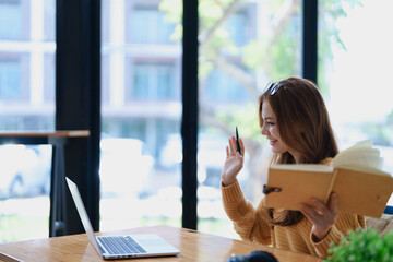 Smiling young woman engaged in a joyful online e-learning video call, gestu while holding a book...