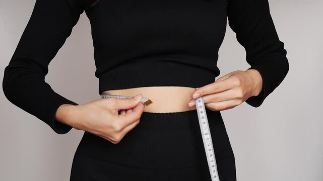 A woman in leggings and a sports top measures her waist and gives a thumbs up.