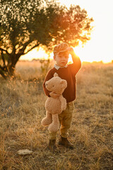 Little boy with a teddy bear walking through an olive grove at sunset. Warm rural scene with yellow grass, soft light, and peaceful childhood atmosphere.