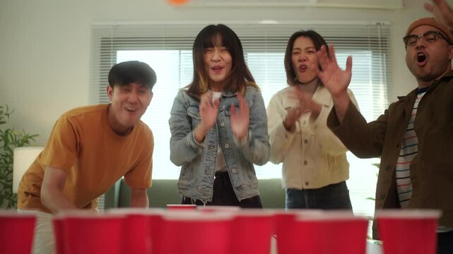 A group of young Asian friends is playing a fun game of ping pong ball toss into red cups at a lively party. The friends enjoy their time together, laughing and competing in the game.
