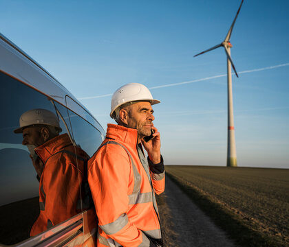 Engineer in high visibility jacket and helmet using cellphone near wind turbine