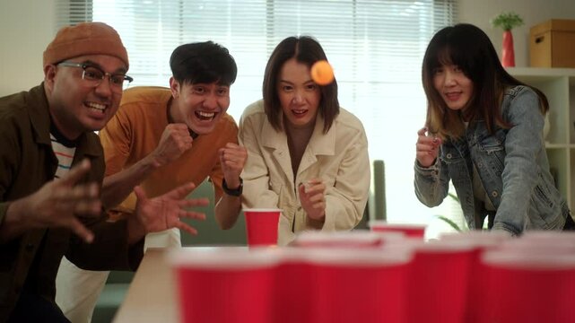 A group of young Asian friends is playing a fun game of ping pong ball toss into red cups at a lively party. The friends enjoy their time together, laughing and competing in the game.
