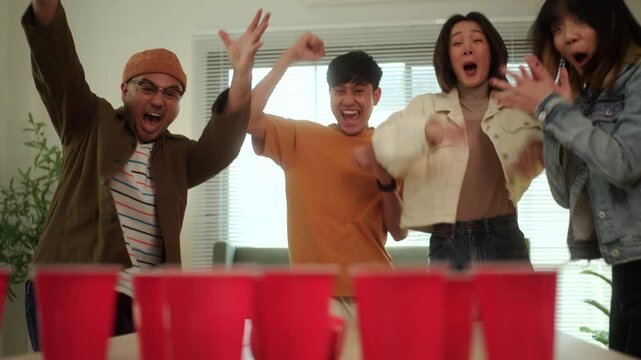 A group of young Asian friends is playing a fun game of ping pong ball toss into red cups at a lively party. The friends enjoy their time together, laughing and competing in the game.
