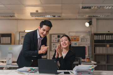 Two happy asian businesspeople are clapping while having a video conference on a tablet in a modern office at night, celebrating a successful project or good news