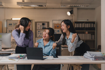Three young asian businesswomen are cheering and raising their fists in celebration of a successful project outcome while working late in the office