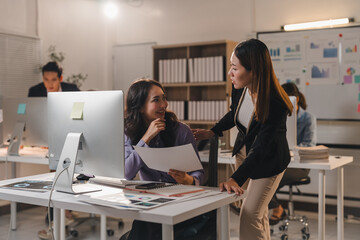 Two young asian businesswomen are discussing over paperwork and working late night together in modern office with desktop computer, using data sheets, graphs, and charts