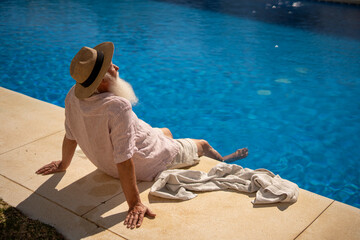 Happy elderly man relaxing by the pool with feet in water, enjoying retirement and sunny leisure lifestyle in Spain on a beautiful summer day