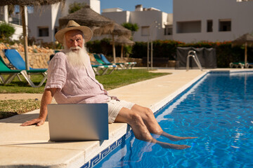 Elderly man enjoying retirement life in Spain, relaxing by the pool with feet in the water and laptop nearby, symbol of digital freedom and peaceful senior lifestyle