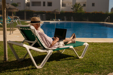 Senior freelancer working remotely on laptop while relaxing on sun lounger by the poolside in sunny resort, enjoying digital nomad lifestyle during retirement