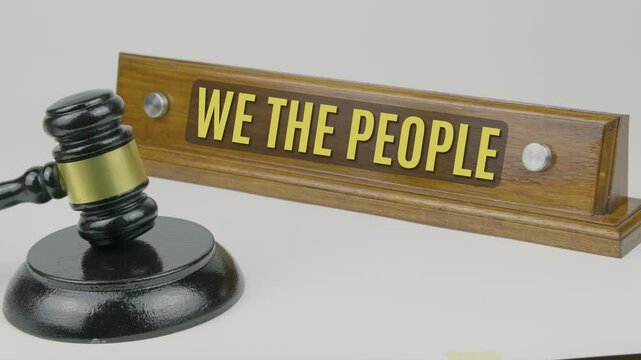 "We the People" text on a wooden nameplate with a gavel tapping once and three times on a white background, symbolizing the preamble of the U.S. Constitution, representing the authority of the people