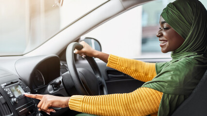 Cheerful African Muslim Woman In Hijab Changing Radio Station While Listening Music In Car,...