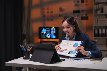 Fototapeta premium Young, cheerful businesswoman presenting charts and graphs during a late-night video call on a tablet, working diligently in a modern office environment