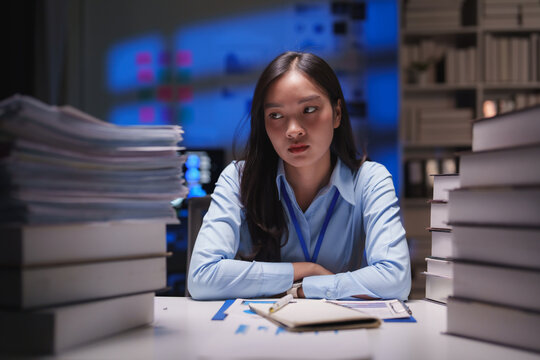 Young businesswoman sitting at her desk late at night, surrounded by piles of paperwork and books, feeling overwhelmed and stressed in a modern corporate office environment