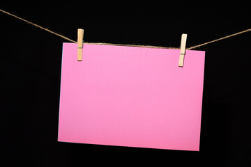 Fotograf&iacute;a de una cartulina rosa sujeta con dos pinzas de madera en una cuerda, con un fondo negro. Ideal para proyectos creativos, dise&ntilde;o gr&aacute;fico y publicidad.