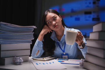 Young office worker holding mug and looking away while sitting at desk with piles of paperwork and books, she is working late at night and feeling tired
