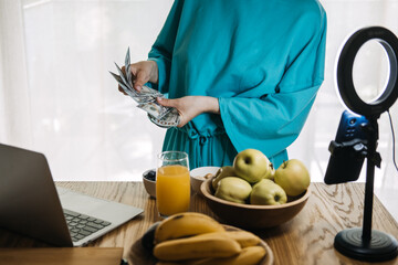 Unrecognizable woman counting US dollar bills beside a laptop, phone on tripod and fruit on table....
