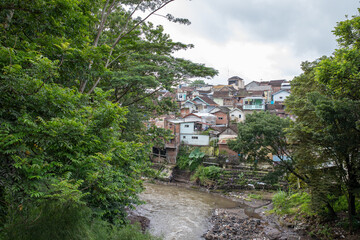 A view of a small river flowing through a green forest with a densely packed residential area in...
