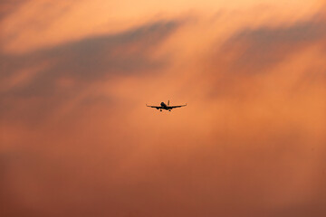 Commercial airplane flying in the distance with a dramatic orange sunset sky, silhouette of aircraft against warm-toned clouds.