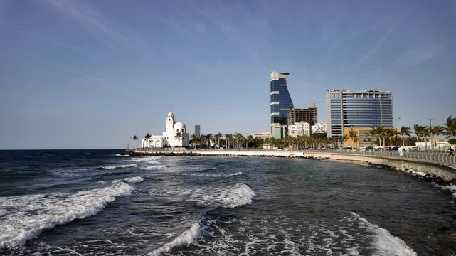 Waves crashing against the shore of the Red Sea with a white mosque and modern buildings rising along the Jeddah Corniche in Saudi Arabia, showcasing a blend of tradition and progress