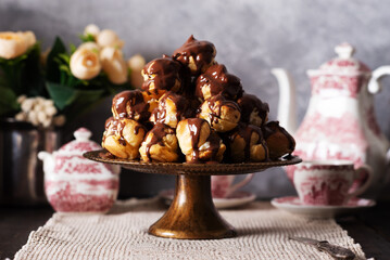 Profiterole cake, or cream puff, is a French choux pastry ball filled with whipped cream, custard, and chocolate sauce, served on a gold plate with cup of tea and teapot.