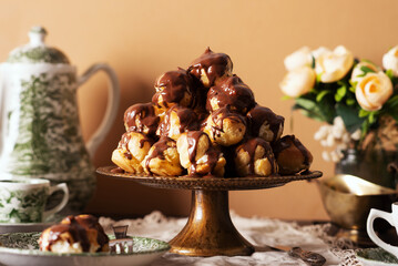 Profiterole cake, or cream puff, is a French choux pastry ball filled with whipped cream, custard, and chocolate sauce, served on a gold plate with cup of tea and teapot.