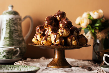 Profiterole cake, or cream puff, is a French choux pastry ball filled with whipped cream, custard, and chocolate sauce, served on a gold plate with cup of tea and teapot.
