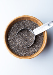 Wooden bowl filled with chia seeds (Salvia hispanica) on a white background.Metal measuring spoon rests inside the bowl, partially submerged in the seeds. 