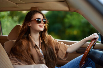 Young woman driving a vintage car in stylish sunglasses, smiling joyfully in a sunny outdoor...