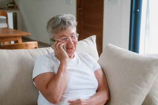 Portrait of senior woman with gray hair sitting on sofa in living room and talking on a smartphone. She shows various emotions and smiles. Grandmother talking to her family Technologies in elderly age