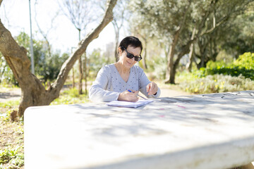 Fototapeta premium Senior woman writing in notebook at park table on sunny day