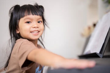 Young Asian girl enjoy plays piano at home with smile and looks at camera. Little kid prcatices music instrument in house. Child interested in musical. © Wanwajee