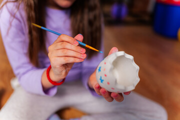 Young girl painting a ceramic easter egg with blue paint, using a small brush to add personal...