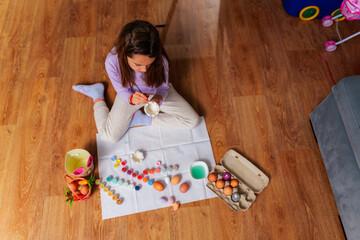Young girl sitting on the floor painting easter eggs at home, surrounded by colorful paints,...