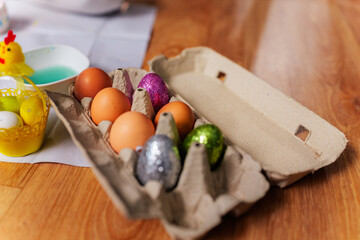 Cardboard egg carton containing natural and decorated easter eggs, with a bowl of blue dye and yellow basket of colorful eggs nearby, suggesting preparations for easter celebration on wooden table