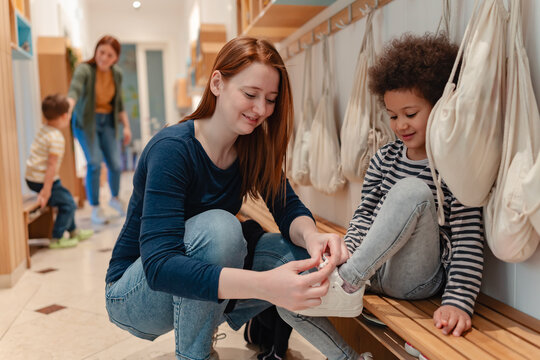 Kindergarten teacher helps a cheerful curly-haired child put on shoes in a bright hallway, sharing a warm and caring moment. - Powered by Adobe