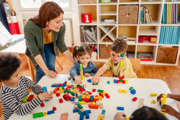 Smiling teacher interacts with diverse group of young children playing with colorful blocks in a bright, cheerful classroom setting.