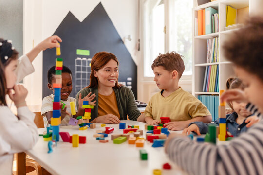 Smiling teacher interacts with diverse group of young children playing with colorful blocks in a bright, cheerful classroom setting.