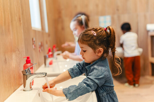 Multicultural preschool children wash their hands at sinks in a wooden-themed bathroom, practicing hygiene routines as part of their daily classroom activities.