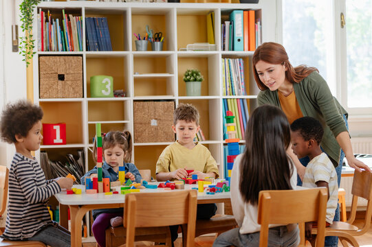Diverse preschool children play with colorful building blocks at a classroom table, guided by a smiling teacher in a creative, inclusive early learning environment.