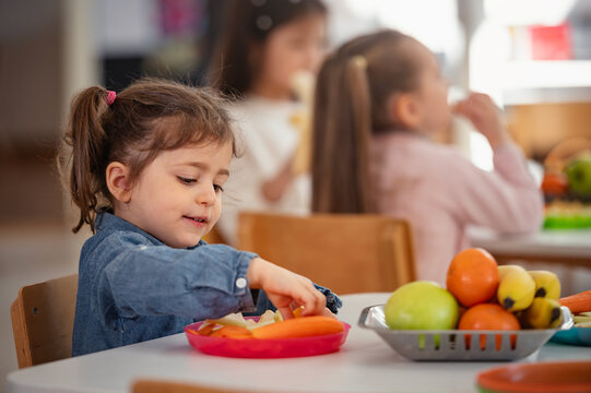 Young girl enjoys a healthy snack at preschool while other children sit nearby, promoting healthy eating habits, nutrition, and diversity in an early childhood classroom setting.