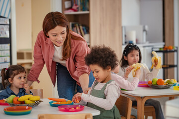 A diverse group of preschool children sits around a table eating healthy snacks while a teacher...
