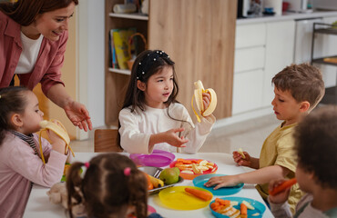 A diverse group of preschool children sits around a table eating healthy snacks while a teacher...