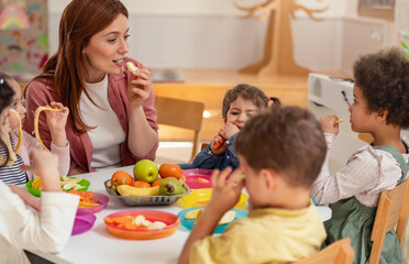 A diverse group of preschool children sits around a table eating healthy snacks while a teacher engages with them, promoting nutrition, inclusion, and social interaction in early education.