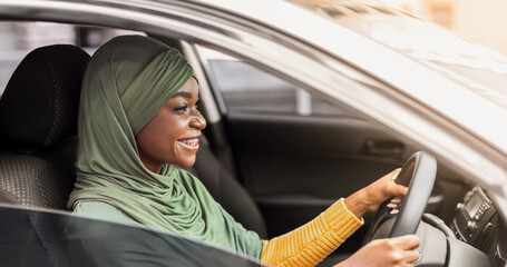Fototapeta premium Cheerful African Muslim Lady In Headscarf Driving New Car In City, Riding Her New Vehicle, Got Driving License, Holding Steering Wheel And Using Seatbelt, Side View Shot With Copy Space