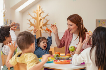 Smiling teacher and diverse group of preschoolers raise their hands and enjoy healthy snacks together, promoting fun, nutrition, and inclusion in a vibrant early learning environment.