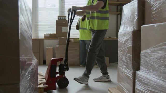 Midsection shot of unknown man moving wooden pallet with heavy boxes wrapped in film using hand truck, female coworker scanning barcodes in background
