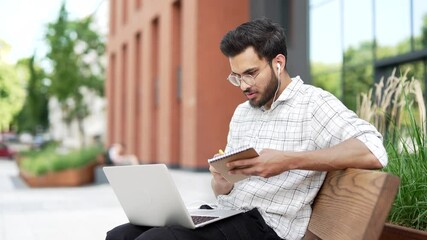 Male student studies online using laptop taking notes while sitting on a bench in campus near university building. Man watching video call training, listens to course. Freelancer communicates remotely - Powered by Adobe