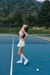 Athletic young woman in a white tennis outfit poses confidently on a court surrounded by green...