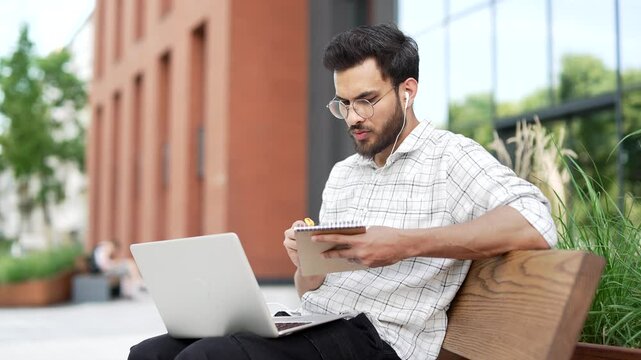 Handsome male student studies online using laptop taking notes while sitting on bench on street near an university building. A man watching video call training, listens remotely to a course or webinar