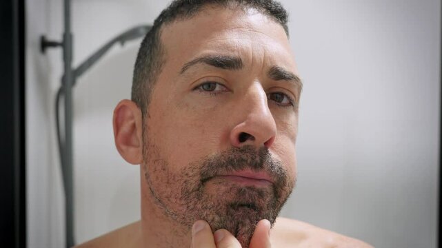 Middle-aged man checking  in slow motion his beard in the mirror, touching face thoughtfully before shaving. 